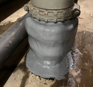 A pipe rising vertically from a concrete floor is repaired at the base, covered with a light gray coating.
