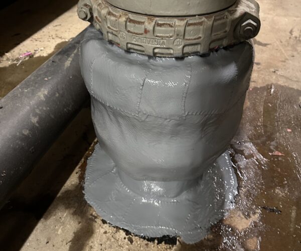 A pipe rising vertically from a concrete floor is repaired at the base, covered with a light gray coating.